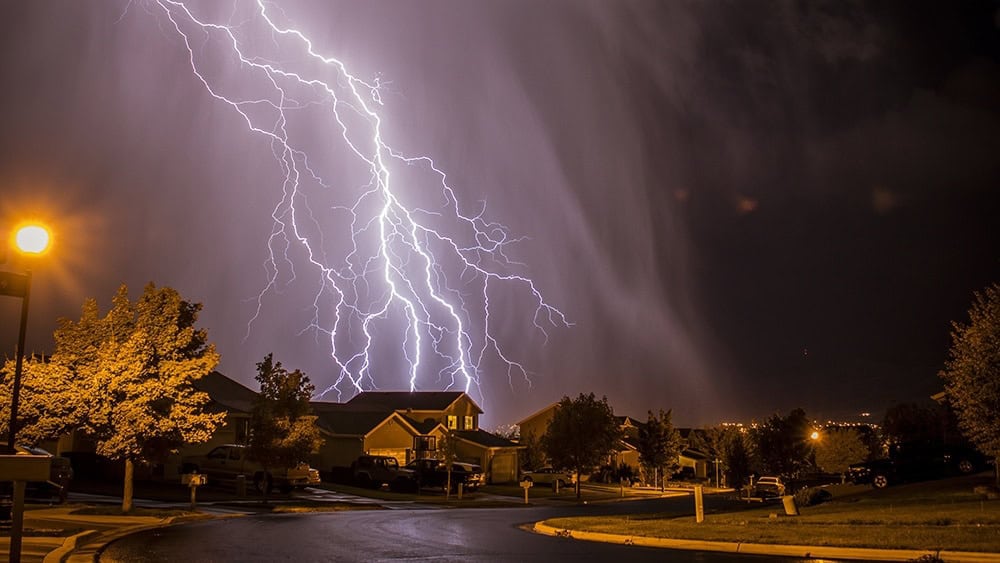 Lightning strikes illuminate a suburban neighborhood at night, highlighting houses and trees under a cloudy sky. Ensure your home deserves the best by investing in whole-home surge protection to keep everything safe during nature's dazzling displays.