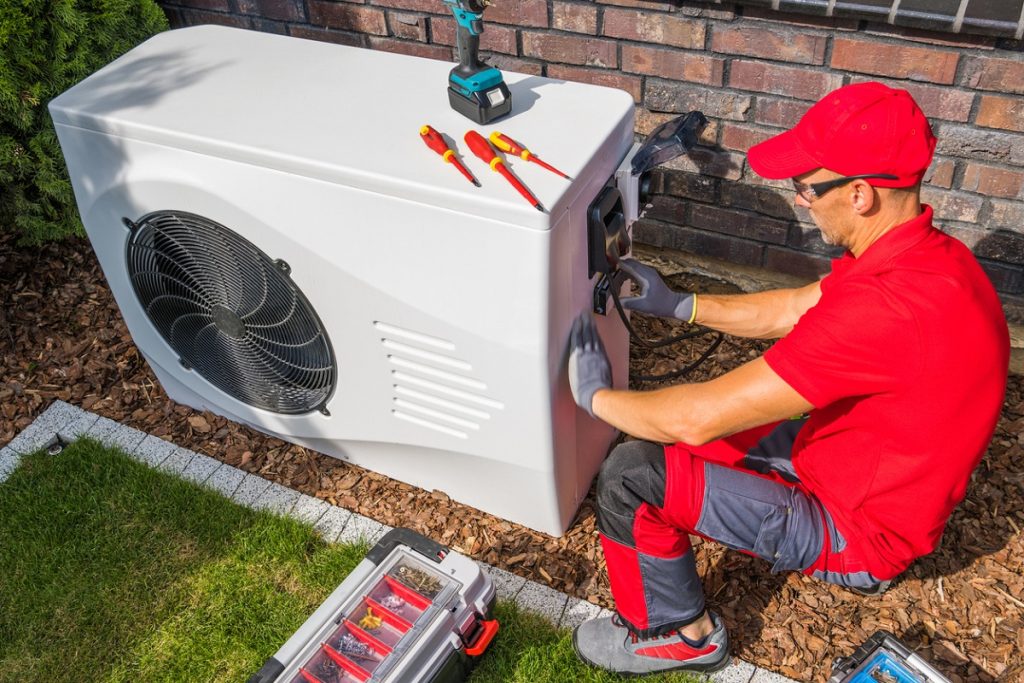 An HVAC technician wearing a red uniform, cap, and protective gloves repairs an outdoor heat pump unit beside a brick wall, with screwdrivers, a power drill, and a toolbox nearby.